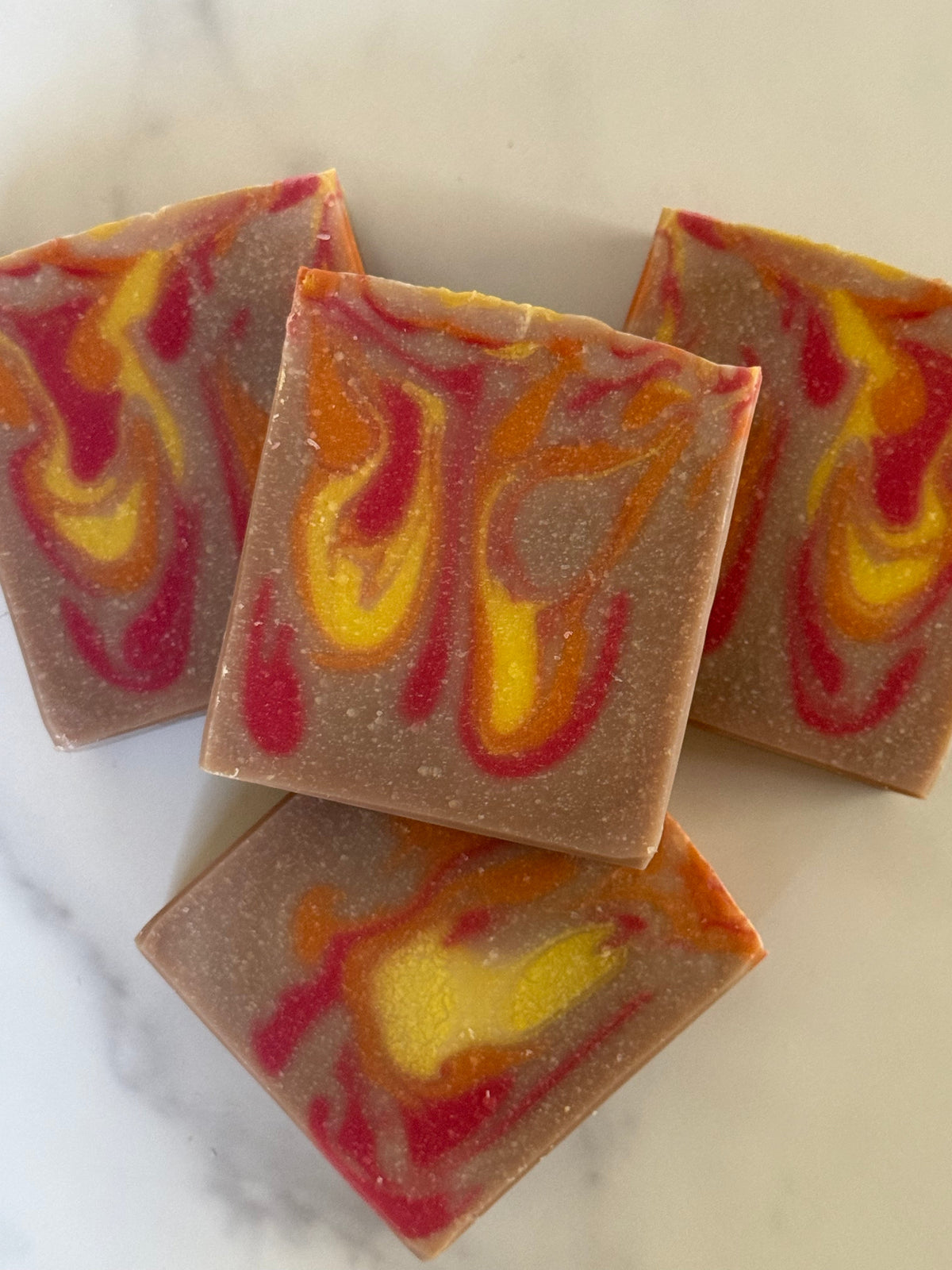 Four square bars of handmade soap with a swirled pattern of orange, yellow, and red on a light beige background, arranged on a marble surface.