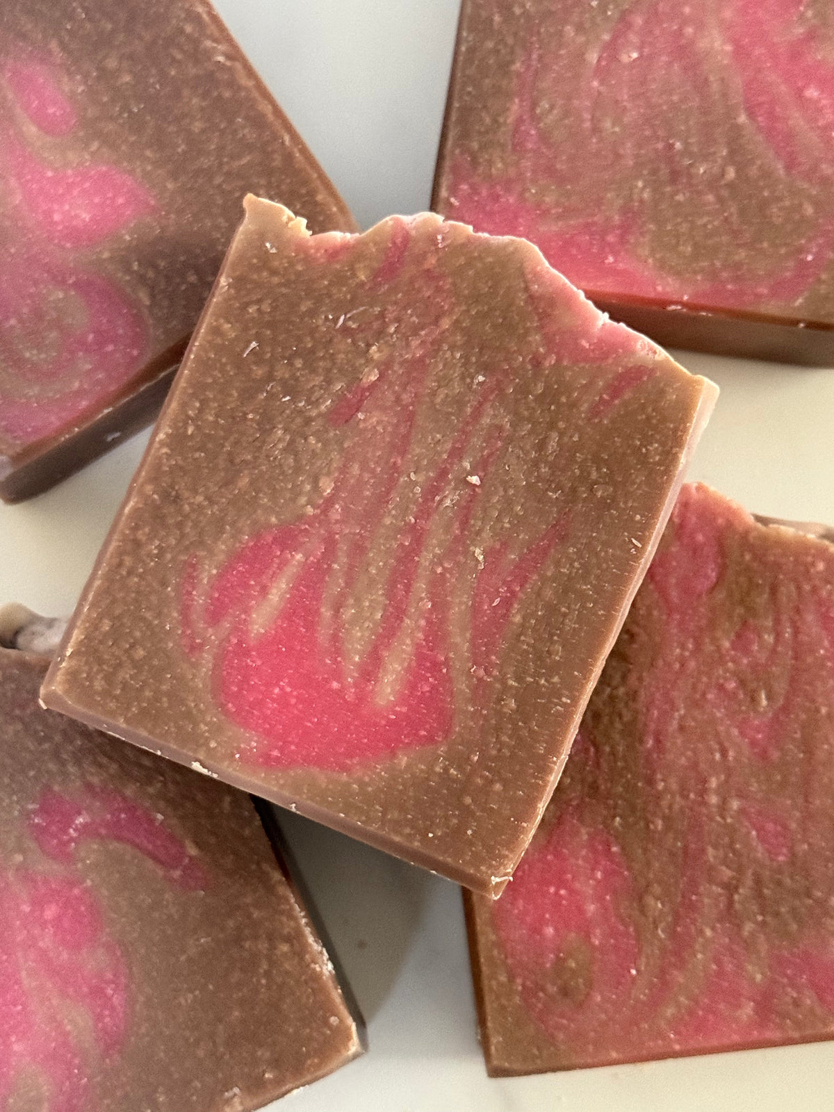 Close-up of several rectangular bars of handmade soap stacked together. The soap has a medium brown base with a pink marbled swirl pattern.