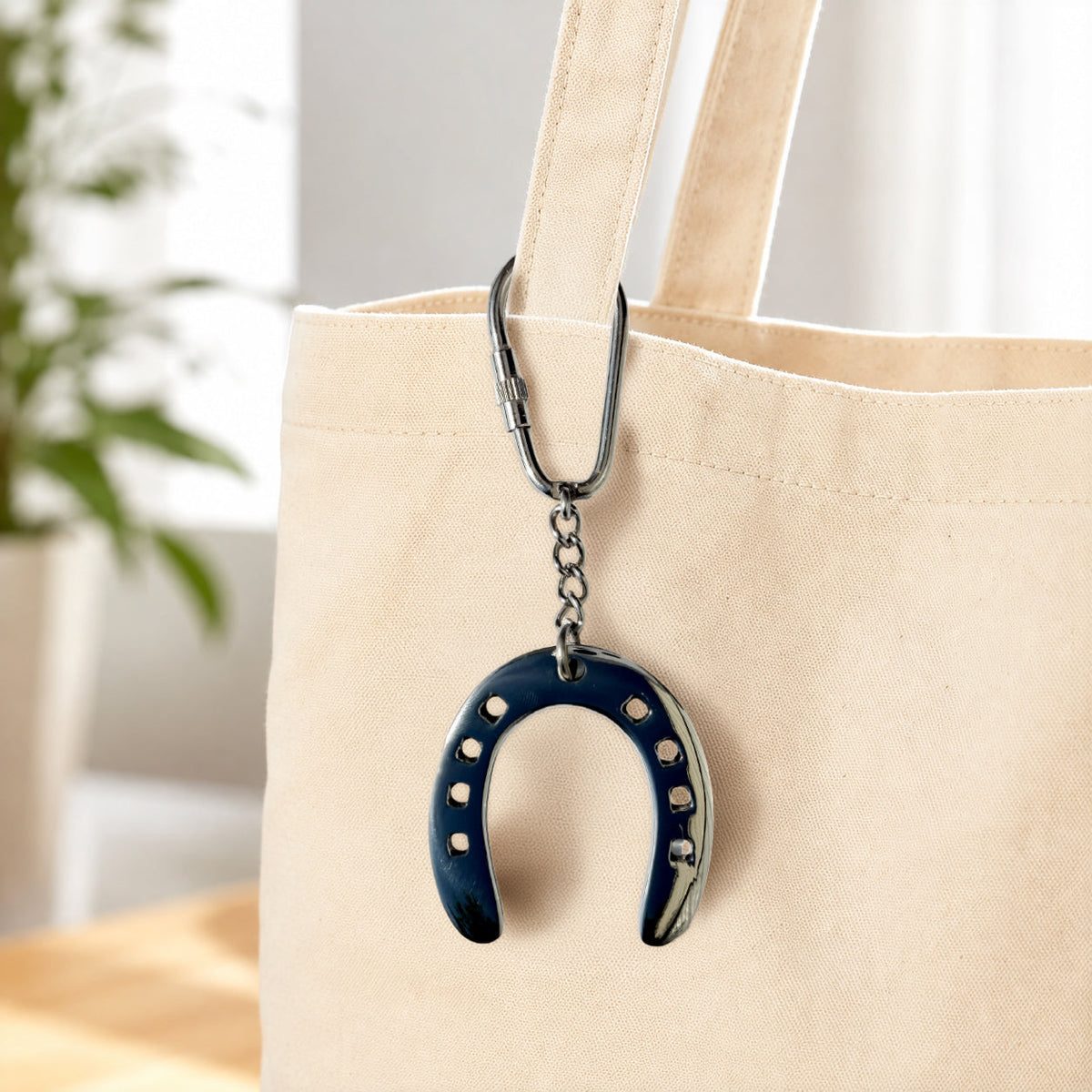 A navy blue horseshoe-shaped keychain with silver studs and a silver carabiner, hanging from the strap of a beige canvas tote bag.
