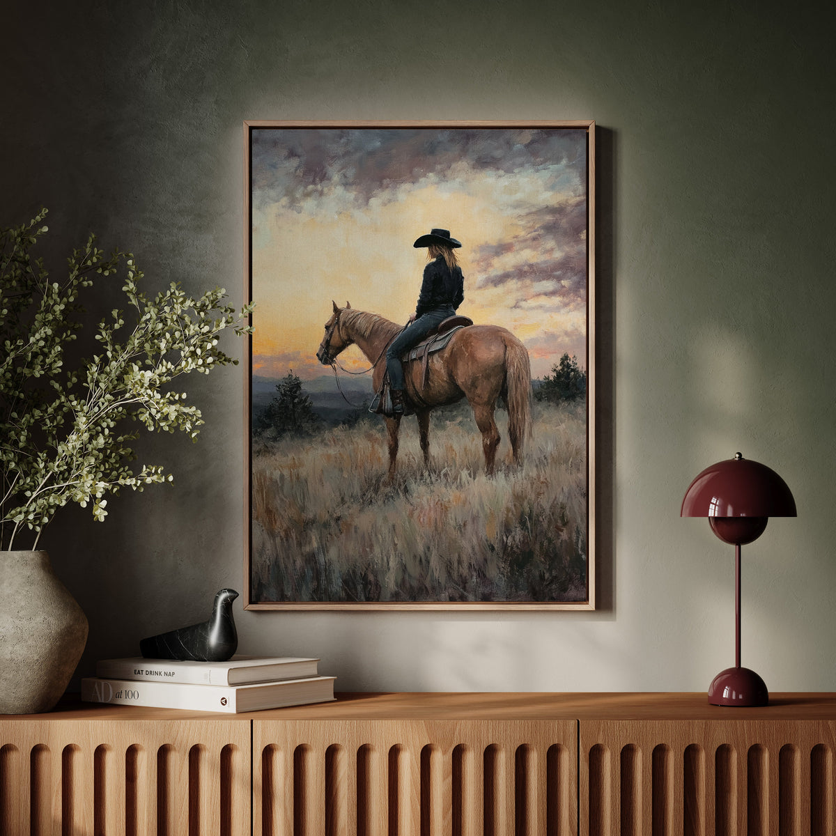 Framed artwork of a person on horseback in a field with a sunset, displayed on a wall above a wooden cabinet.