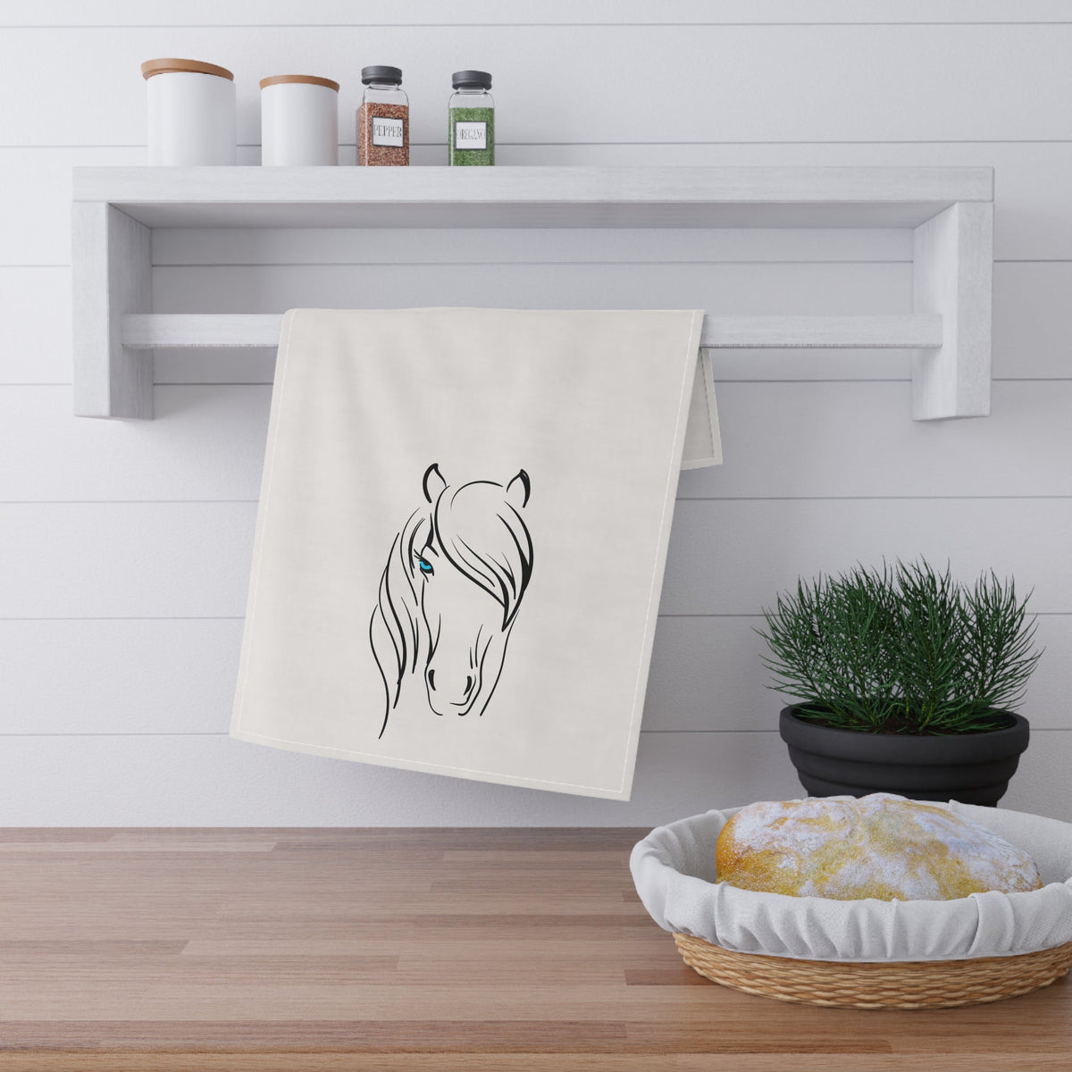 Kitchen towel with horse design hanging on a wooden shelf with a plant and basket below.