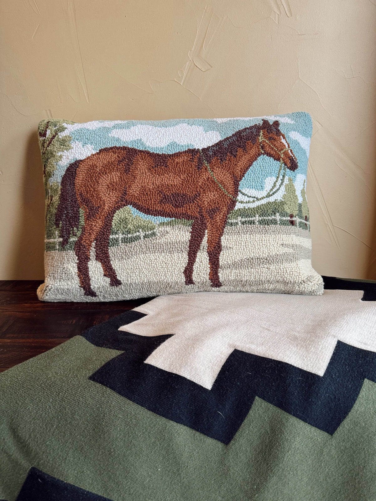 A rectangular needlepoint pillow featuring a brown horse in a field under a blue sky, displayed on a dark wooden surface against a beige wall, with a green and white chevron blanket in the foreground.