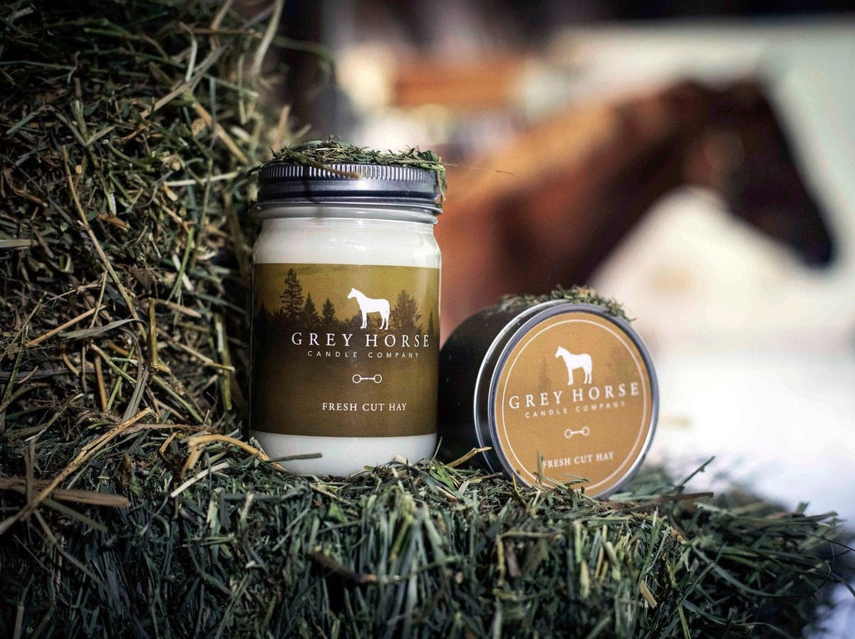 Two candles from Grey Horse Candle Company on a bed of hay with a blurred background.