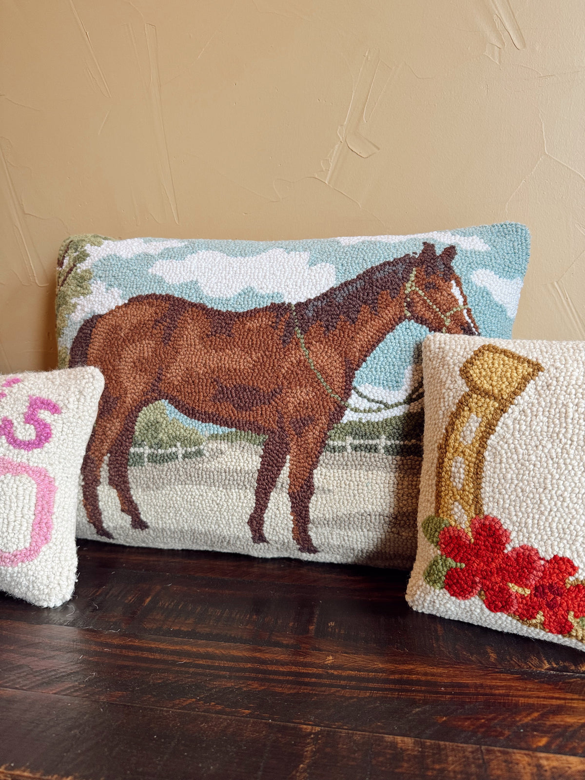 A rectangular woven throw pillow featuring a brown horse on a light blue and white sky background, styled on a dark wooden table with two smaller decorative pillows.