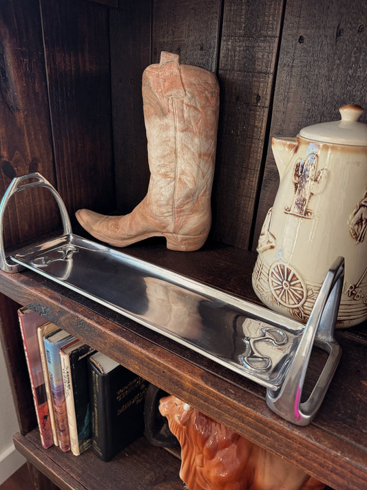A polished stainless steel serving tray with decorative handles, displayed on a dark wooden shelf next to a cowboy boot and a ceramic teapot.