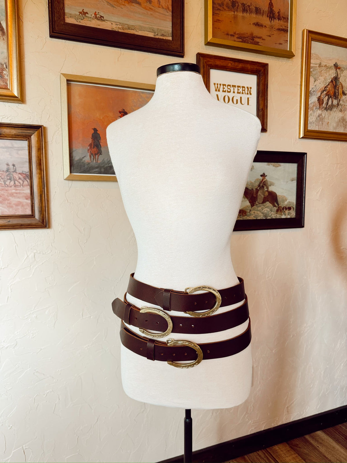 Mannequin wearing a belt with multiple buckles against a wall with framed pictures.
