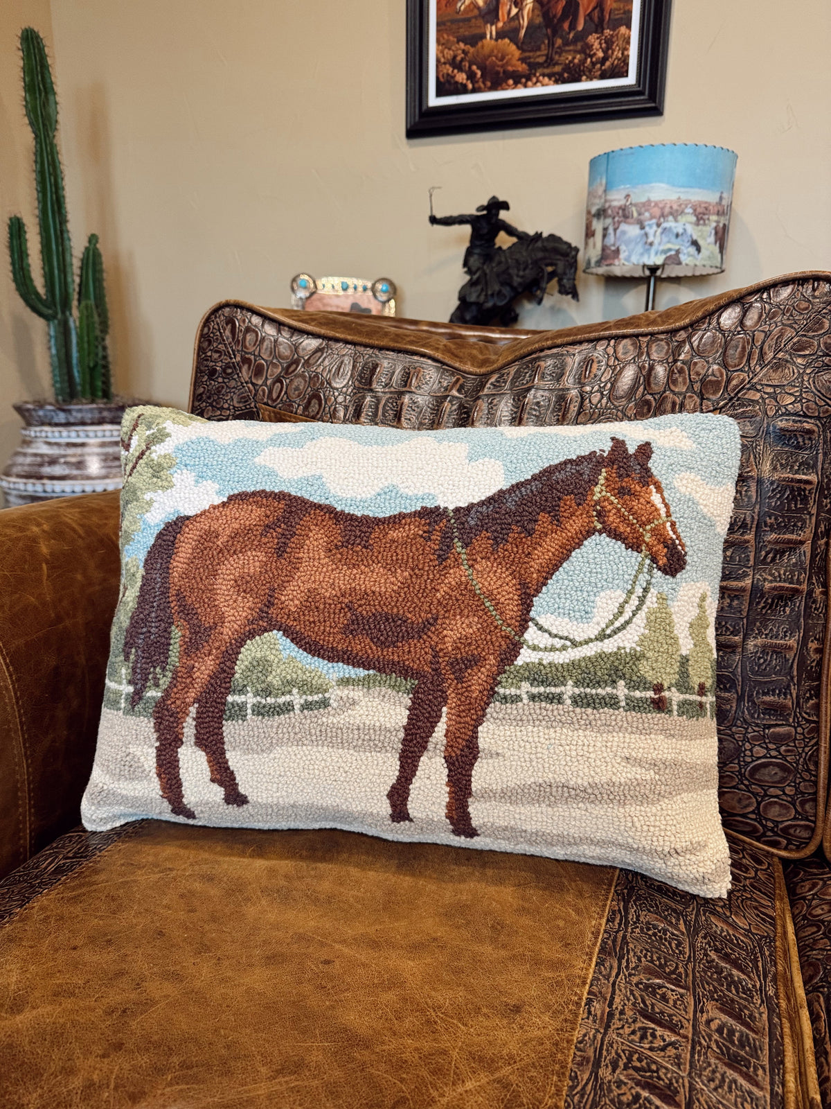 A rectangular woven throw pillow featuring a brown horse illustration, resting on a brown leather and embossed leather armchair in a living room setting.