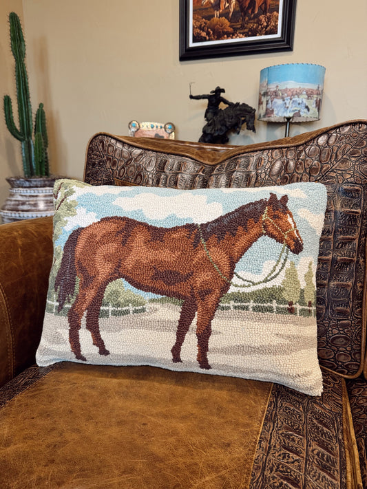 A rectangular woven throw pillow featuring a brown horse illustration, resting on a brown leather and embossed leather armchair in a living room setting.