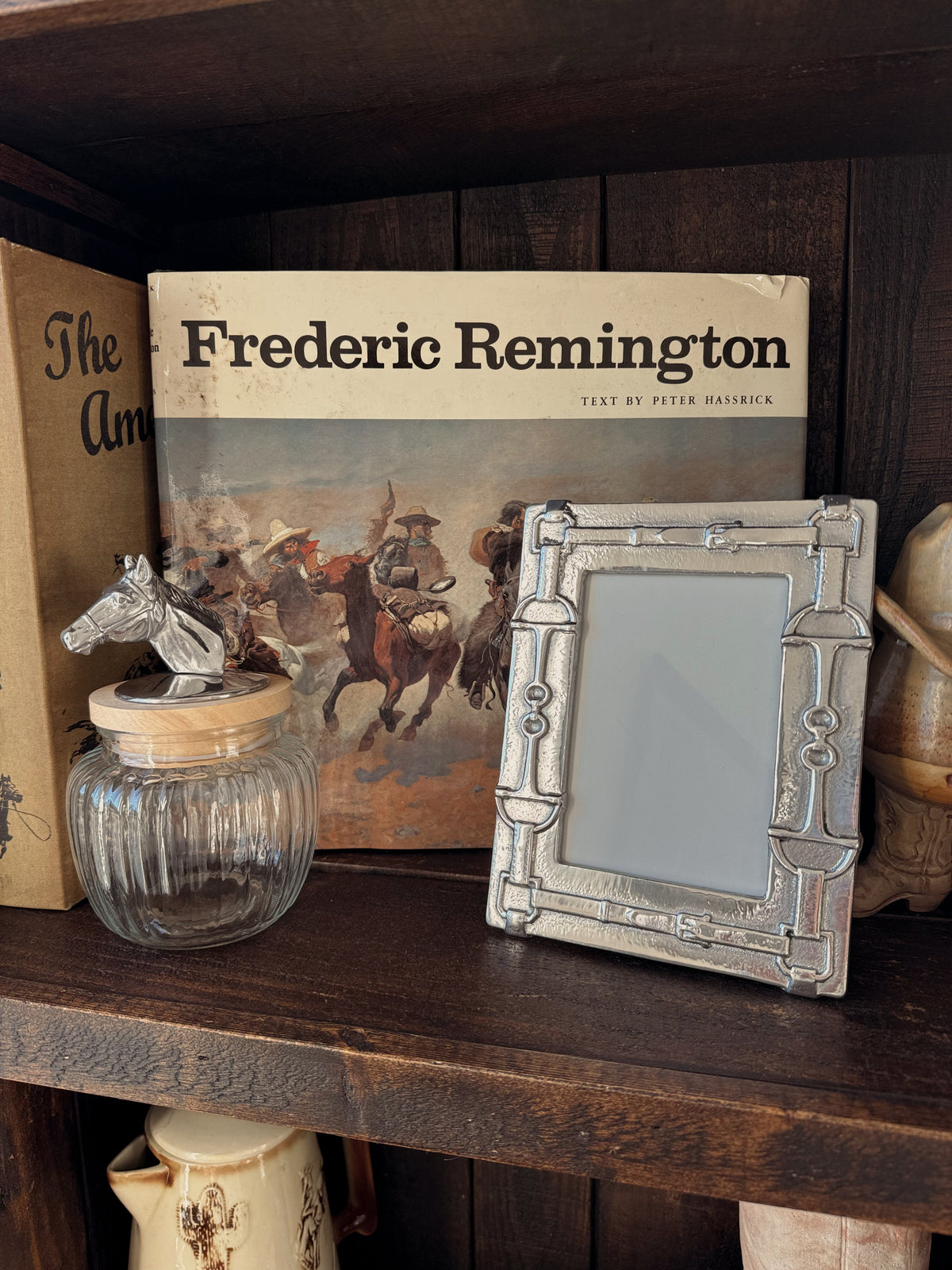 Decorative items on a wooden shelf including a book titled 'Frederic Remington', a picture frame, and a glass jar.