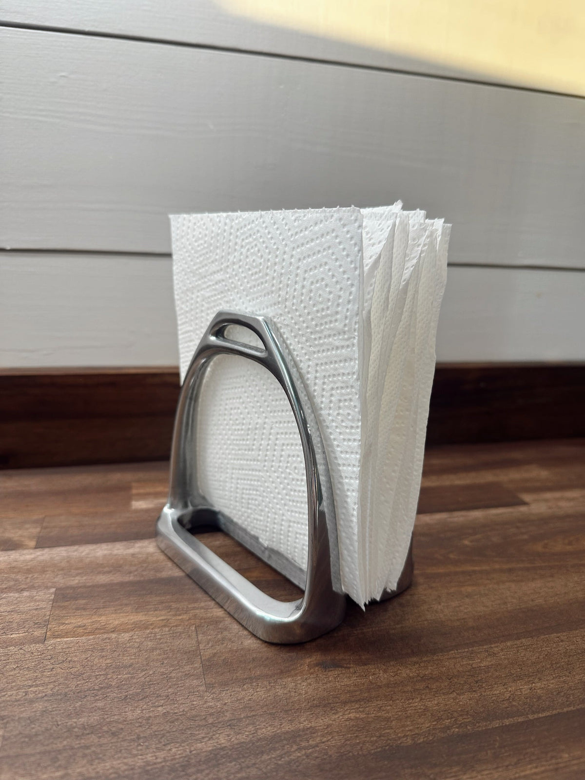 Metal stirrup-shaped napkin holder with white paper towels on a wooden surface.