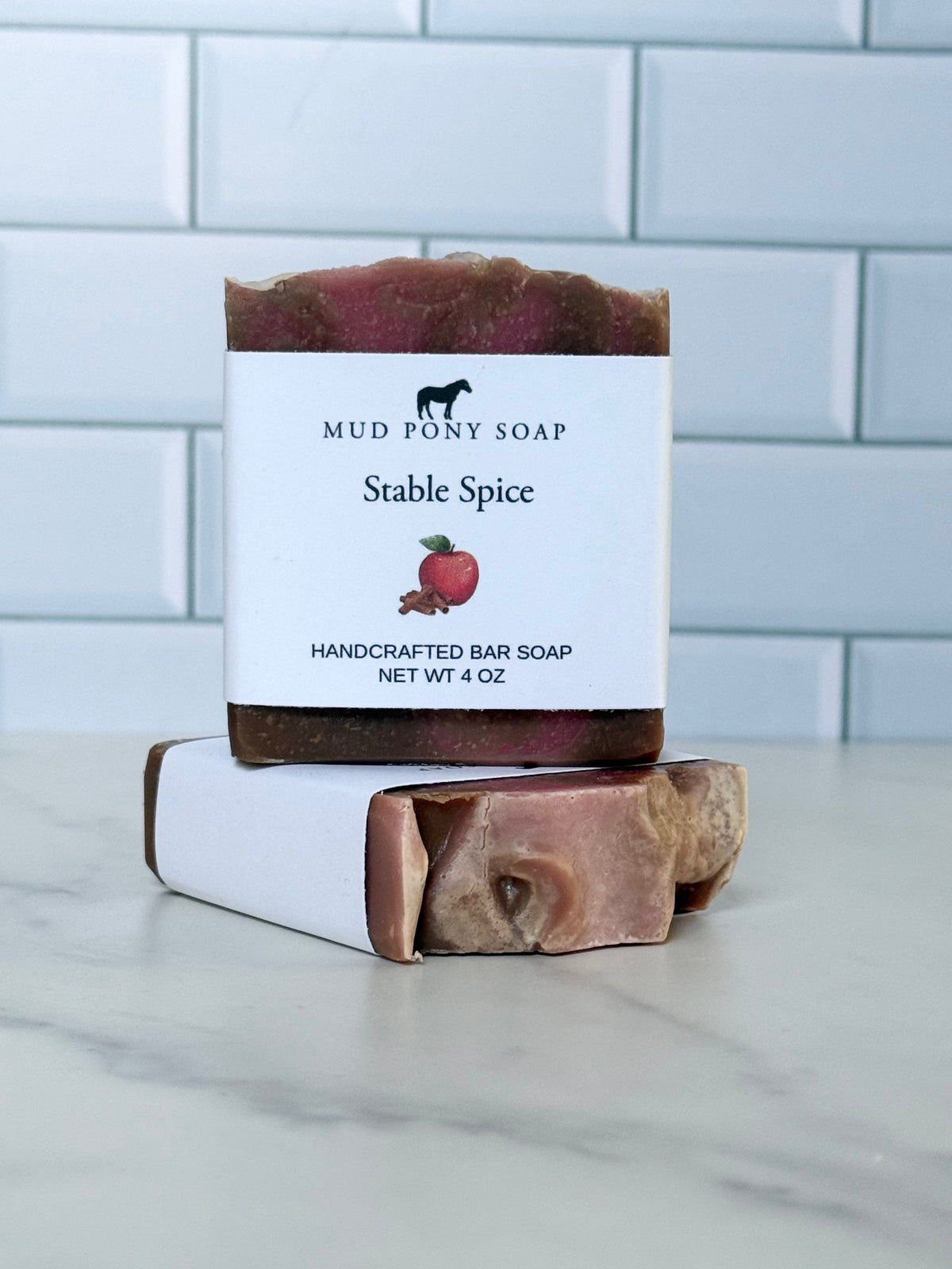 A handcrafted bar of soap labeled 'Mud Pony Soap Stable Spice' stacked on top of another soap bar, set on a light marble countertop with a white subway tile backsplash.