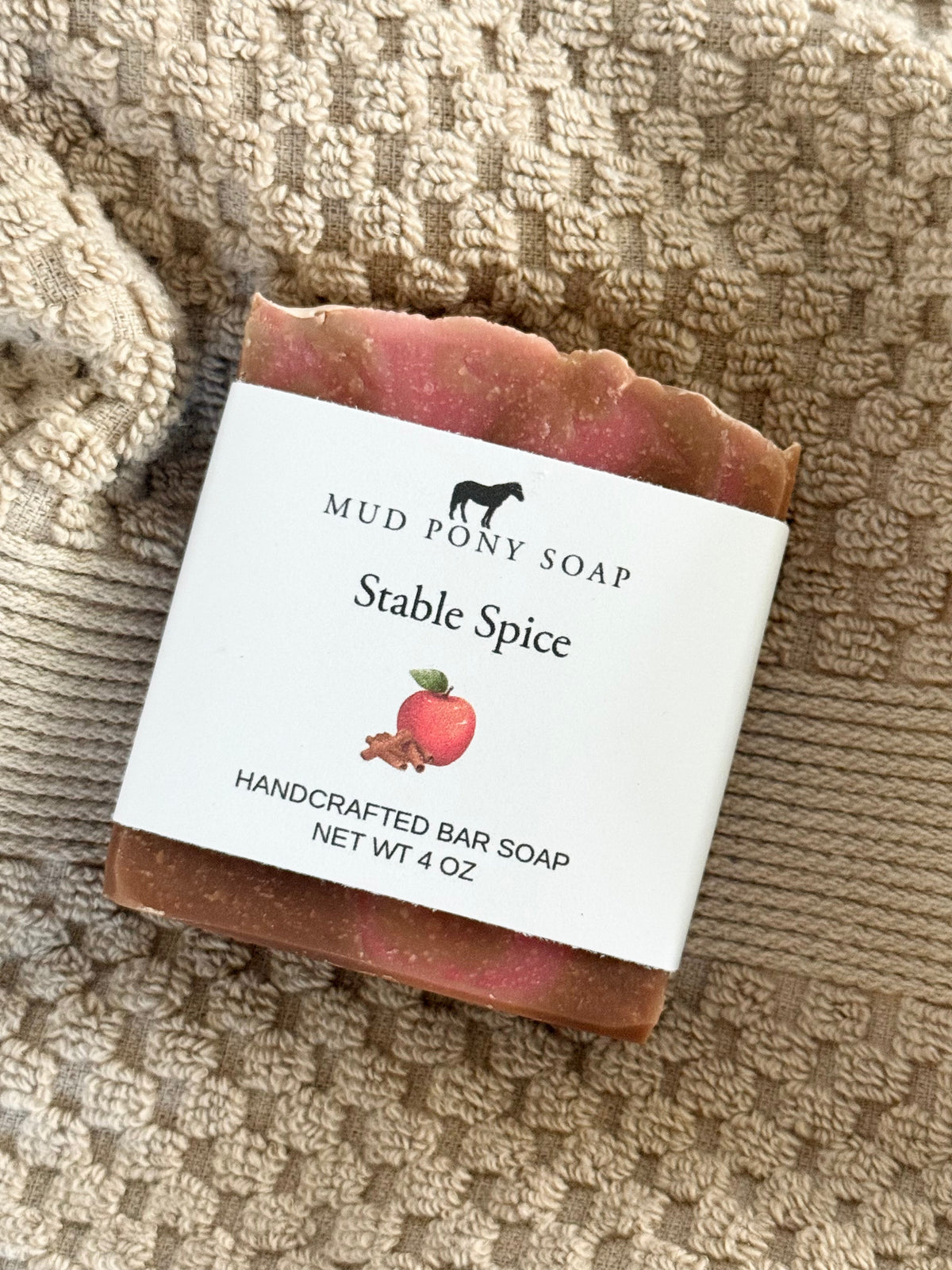 A handmade bar of soap labeled 'Mud Pony Soap Stable Spice' resting on a textured beige knit blanket. The soap has a rustic, scalloped edge and a dusty rose-brown color, with a white paper label showing a small illustration of an apple and cinnamon sticks.