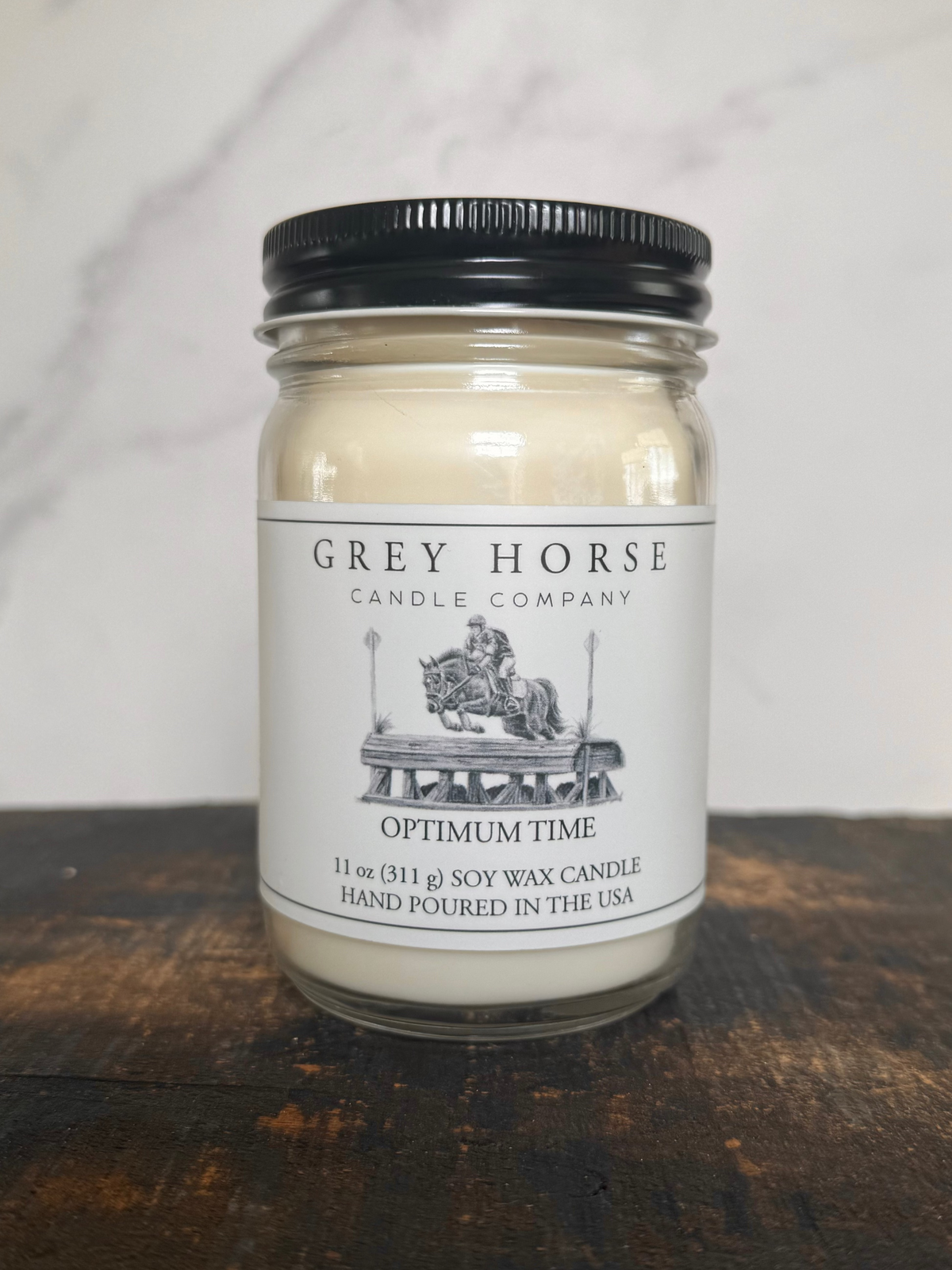 A 11 oz soy wax candle in a clear glass jar with a black metal lid and a white paper label reading 'GREY HORSE CANDLE COMPANY' and 'OPTIMUM TIME'. The candle is placed on a dark wooden surface against a light marble-patterned background.