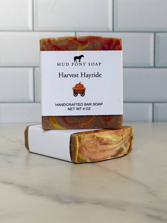 A handcrafted bar soap labeled 'Mud Pony Soap Harvest Hayride' stands upright on a marble-like surface with another wrapped bar resting in front, against a white subway tile background.