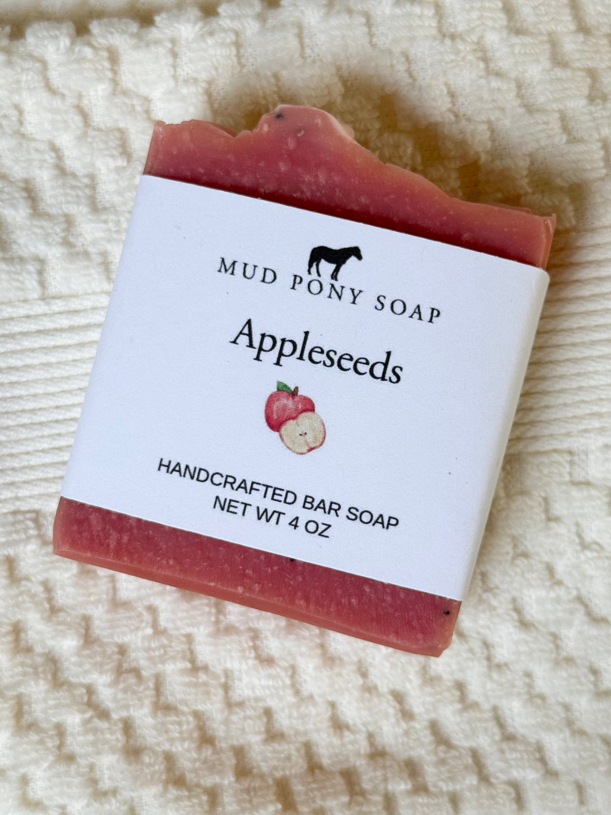 A handcrafted bar of appleseed soap by Mud Pony Soap, wrapped in a white paper label, resting on a textured cream-colored towel.