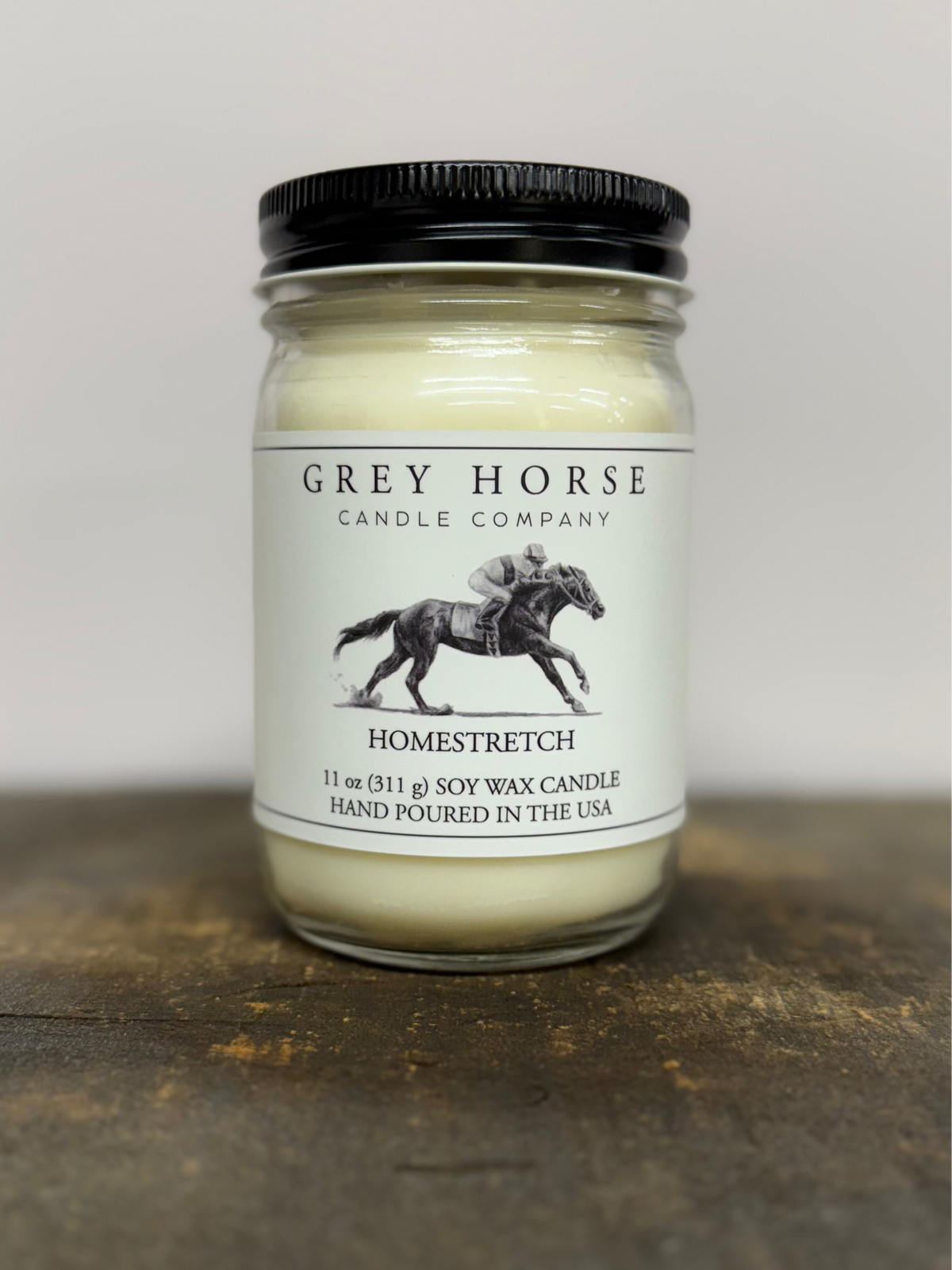 A 11 oz soy wax candle by Grey Horse Candle Company in a clear glass jar with a black lid and a white label featuring a horse-and-rider illustration. The candle is photographed on a rustic wooden surface against a plain light background.