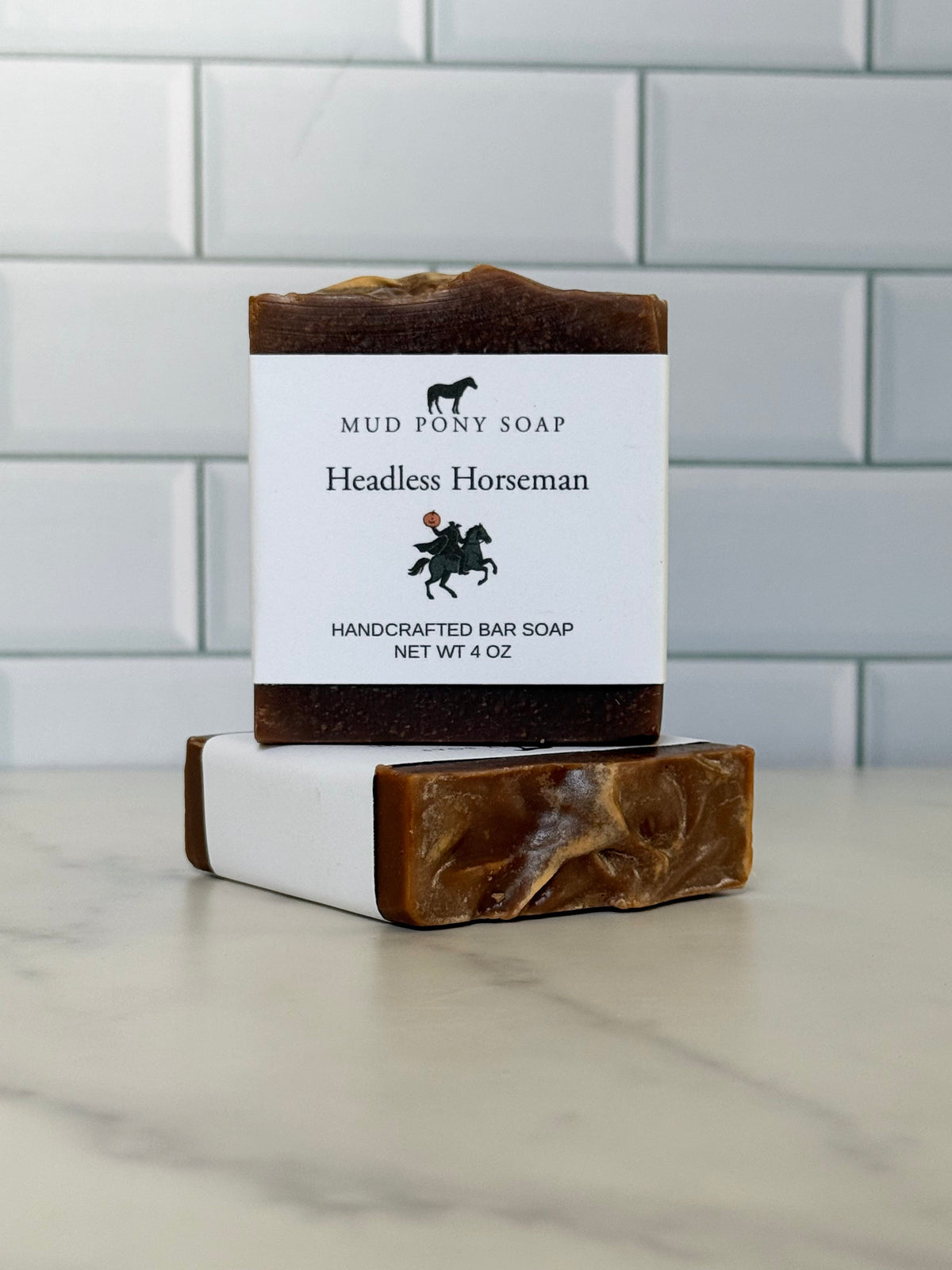 Two handmade bar soaps stacked on a marble countertop in front of a white subway tile wall. The top soap is unwrapped, showing a dark brown marbled bar with a white paper label that reads 'MUD PONY SOAP Headless Horseman HANDCRAFTED BAR SOAP'.