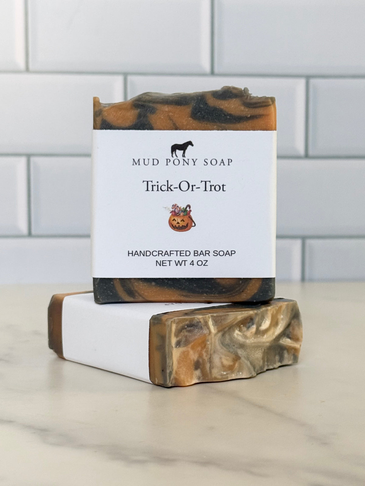 Two handmade bar soaps stacked on a marble surface with a white subway tile background. The top soap is upright with a white paper label reading 'MUD PONY SOAP Trick-Or-Trot HANDCRAFTED BAR SOAP NET WT 4 OZ'.