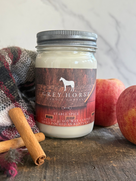 A jar candle labeled Grey Horse Candle Company, Stable Spice, set on a rustic wooden surface with apples, cinnamon sticks, and a plaid blanket in the background.