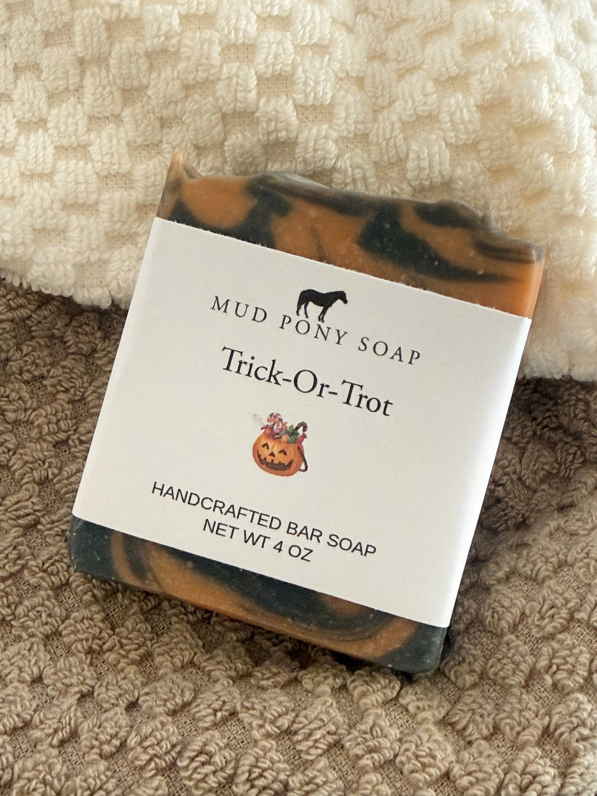 A bar of Mud Pony Soap labeled 'Trick-Or-Trot' resting on a textured beige blanket. The soap has a marbled pattern of orange, black, and brown, with a white paper label showing the brand name and product details.