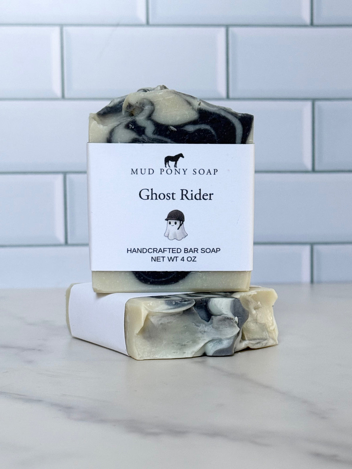 A handcrafted bar of soap with a black, white, and beige marbled pattern, wrapped in a white paper band labeled 'Mud Pony Soap Ghost Rider'. A second unwrapped bar is stacked on top of the wrapped one, set on a white marble surface with a white subway tile background.