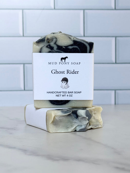 A handcrafted bar of soap with a black, white, and beige marbled pattern, wrapped in a white paper band labeled 'Mud Pony Soap Ghost Rider'. A second unwrapped bar is stacked on top of the wrapped one, set on a white marble surface with a white subway tile background.