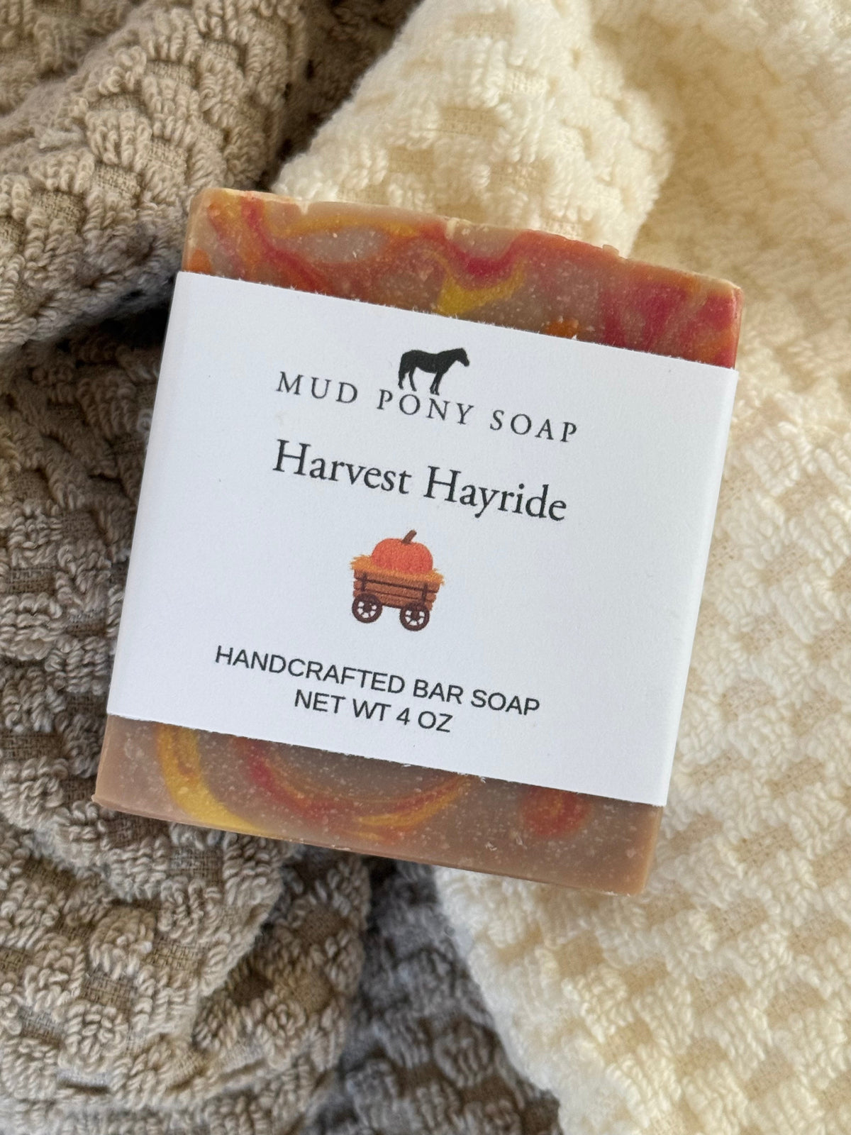 A handcrafted bar of Mud Pony Soap named 'Harvest Hayride' resting on a textured beige and cream blanket. The soap has a marbled pattern of orange, yellow, and brown, with a white paper label wrapped around it.