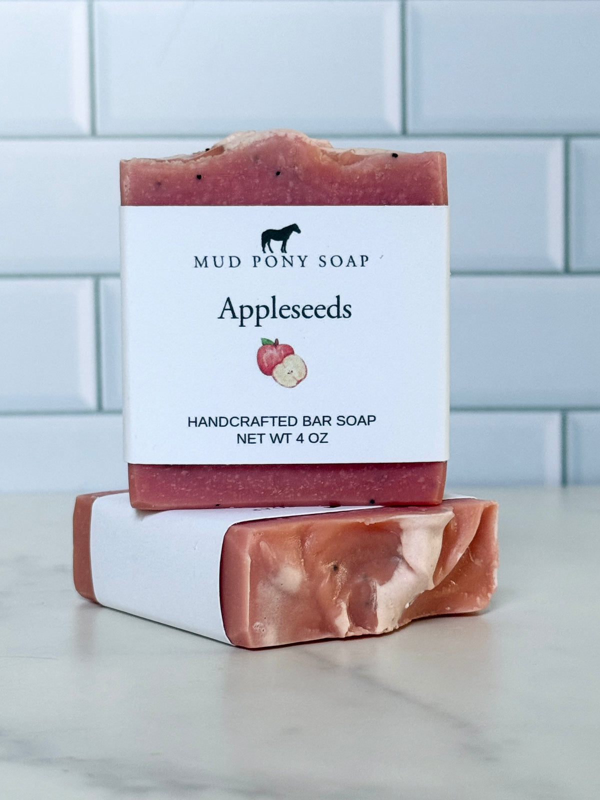 Two bars of Mud Pony Soap 'Applesseeds' handcrafted bar soap stacked on a marble surface with a white subway tile background. The top bar is standing upright with its white paper label showing a black horse logo and apple graphic, while the bottom bar lies flat showing the marbled pink and cream soap texture.