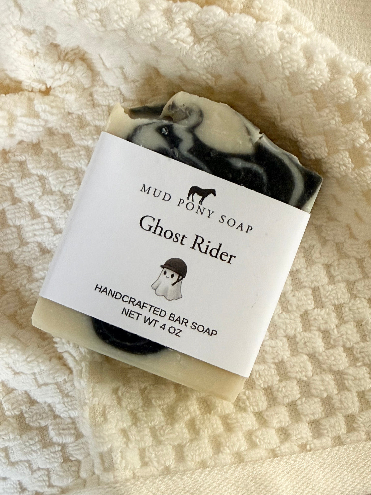 A 4 oz bar of Mud Pony Soap called 'Ghost Rider' resting on a textured cream-colored blanket. The soap has a marbled black and cream-colored pattern, and it is wrapped in a white paper label with black text and illustrations.