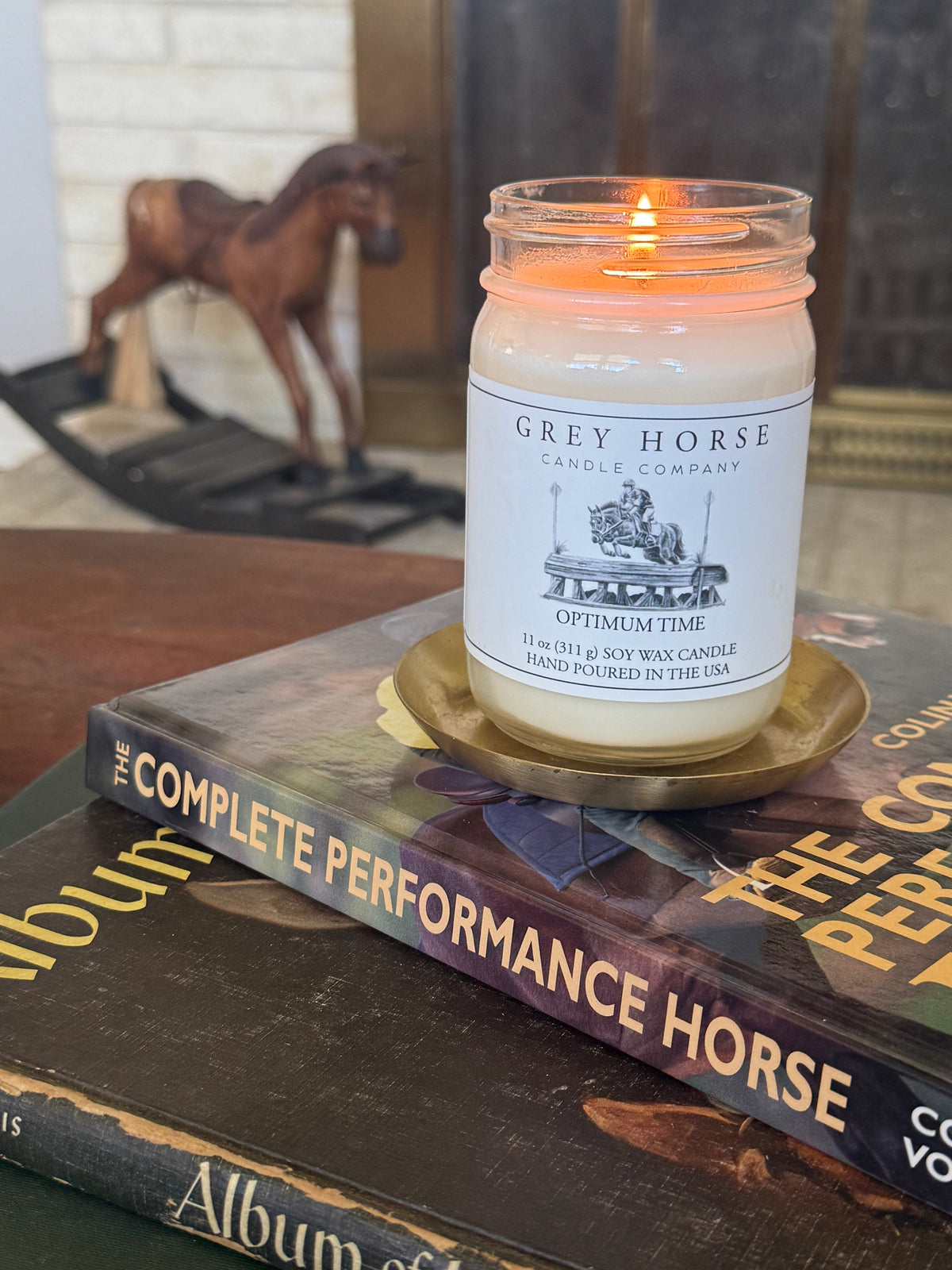 Candle in a jar labeled 'Grey Horse Candle Company' on top of books with a horse model in the background.
