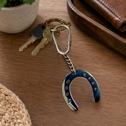 A horseshoe-shaped keychain with a dark blue inlay and silver metal edges, attached to a split key ring with two keys, displayed on a wooden tabletop with a leather wallet, plant pot, and woven mat in the background.