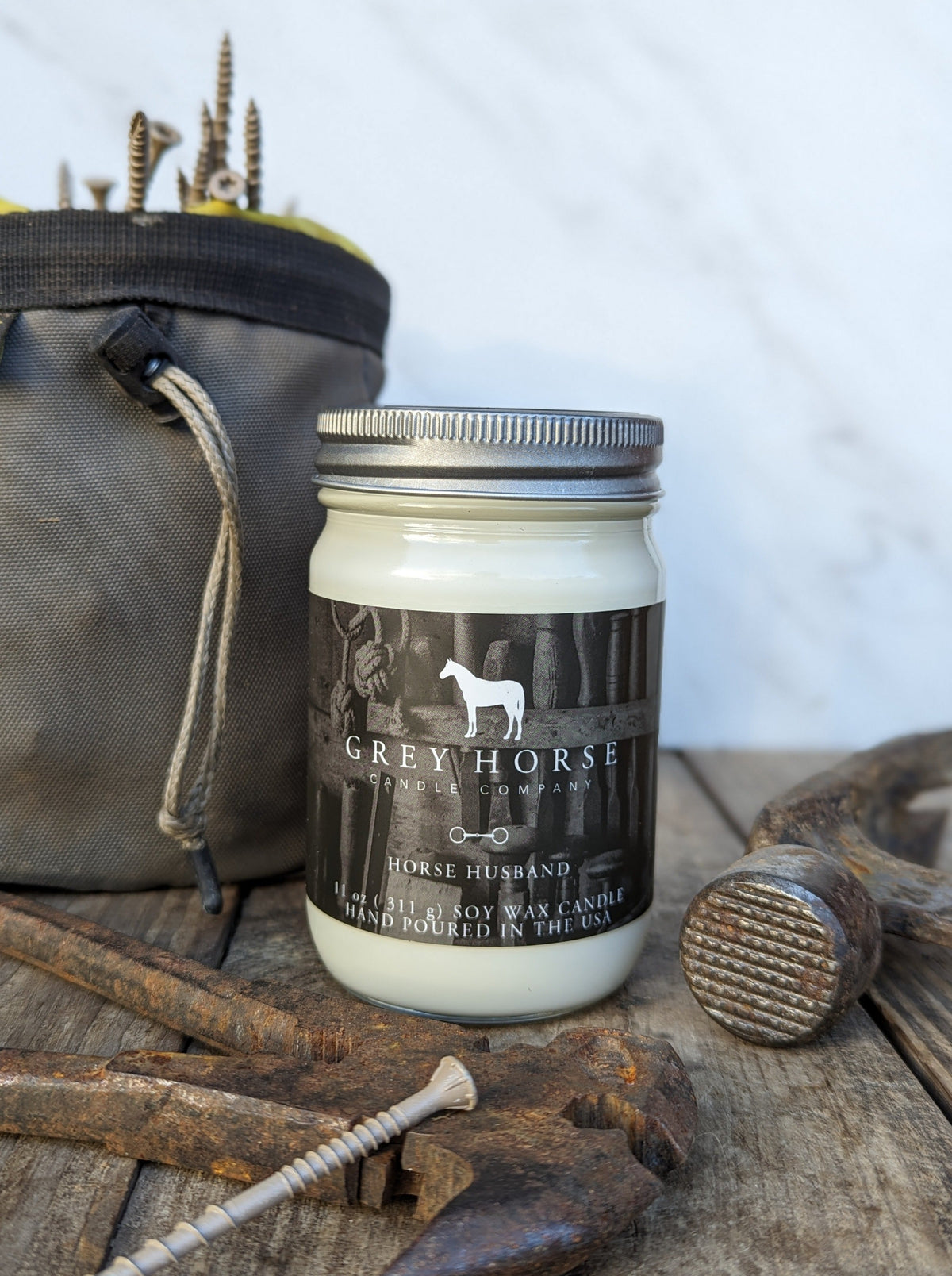 A jar candle labeled 'Grey Horse Candle Company' and 'Horse Husband' sits on a rustic wooden surface surrounded by horse-themed props, including a mallet, nails, and a canvas bag.