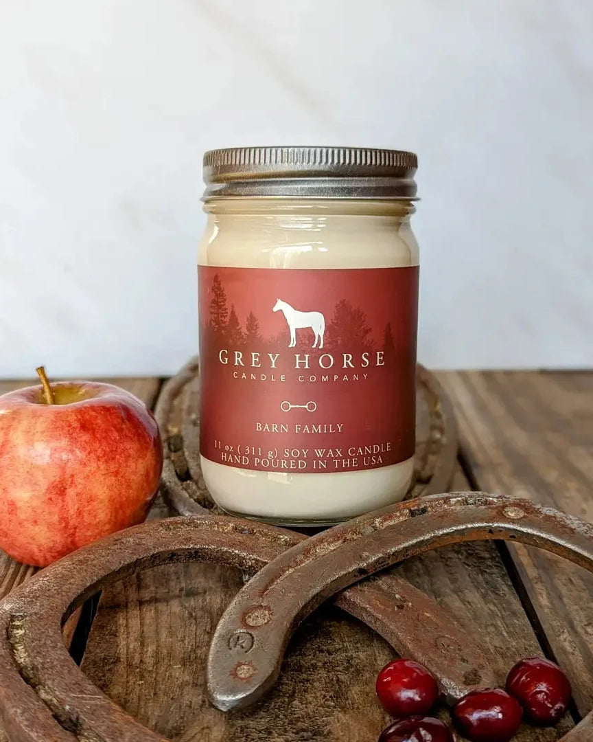 A 11 oz scented candle in a clear glass mason jar with a metal screw-top lid and a dark red label reading 'GREY HORSE CANDLE COMPANY' and 'BARN FAMILY.' The jar is styled on a rustic wooden surface with an apple, horseshoes, and cranberries around it.