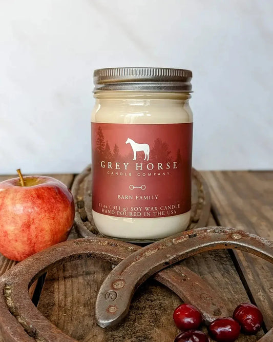 A 11 oz scented candle in a clear glass mason jar with a metal screw-top lid and a dark red label reading 'GREY HORSE CANDLE COMPANY' and 'BARN FAMILY.' The jar is styled on a rustic wooden surface with an apple, horseshoes, and cranberries around it.
