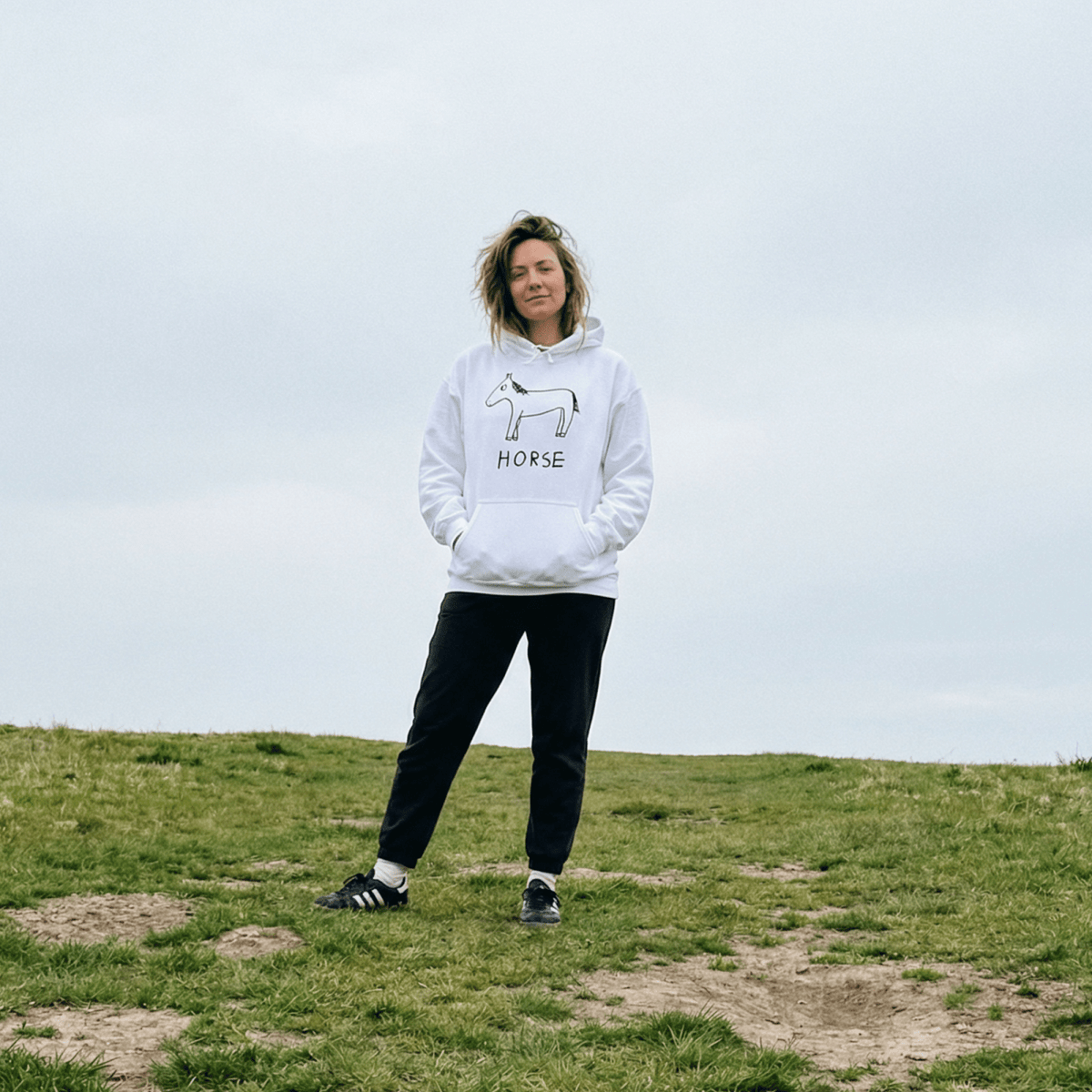 Person wearing a white hoodie with 'Horse' text and a horse graphic on a grassy hill.