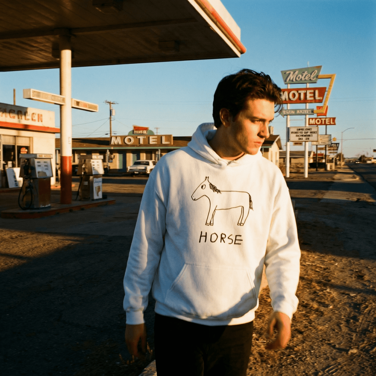 Man wearing a hoodie with a horse graphic in front of a gas station and motel signs.