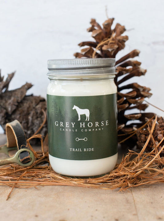 A jar candle from Grey Horse Candle Company labeled 'Trail Ride' sits on a bed of dried grass with pinecones and rustic twine in the background. The candle is in a clear glass jar with white wax and a dark green label featuring a white horse silhouette.