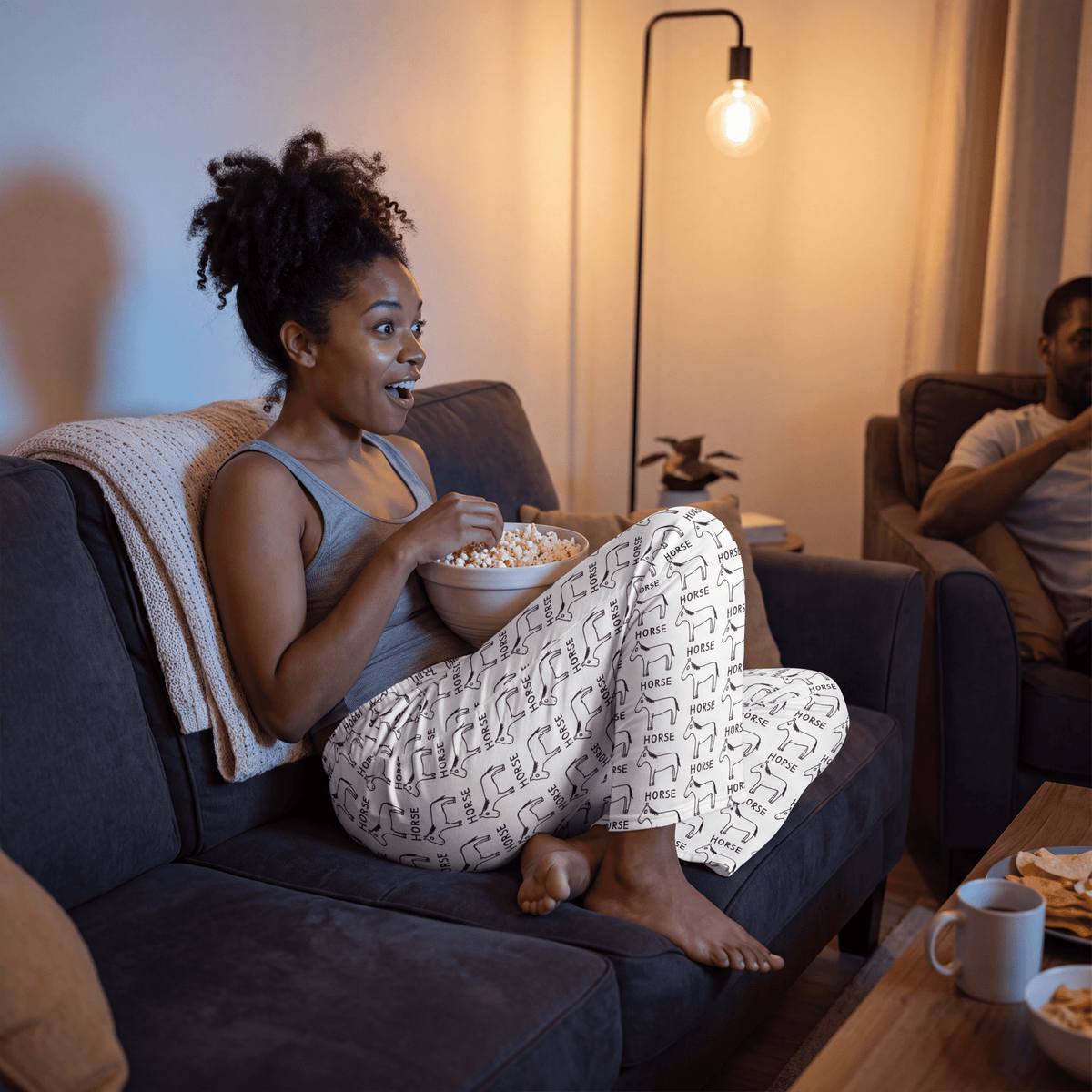 A woman sits on a dark gray couch in a cozy living room, eating popcorn from a bowl. She is wearing a gray tank top and white pajama pants with a black geometric pattern.