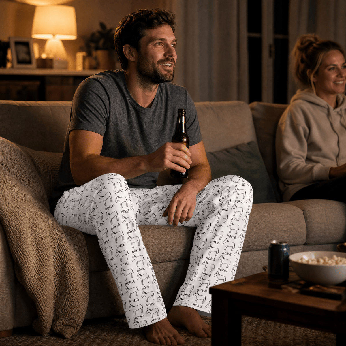 A man sits on a beige couch in a living room, wearing white pajama pants with a black geometric pattern and a dark gray t-shirt, holding a beer bottle. A woman is seated on the couch behind him, and a coffee table with popcorn and a soda can is in the foreground.
