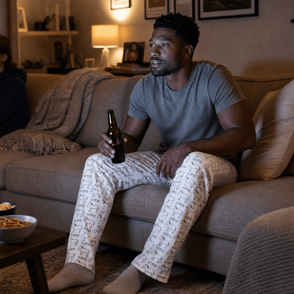 A man sits on a beige couch in a warmly lit living room, holding a brown glass beer bottle. He is wearing a gray t-shirt and cream-colored pajama pants with a repeating black script pattern.