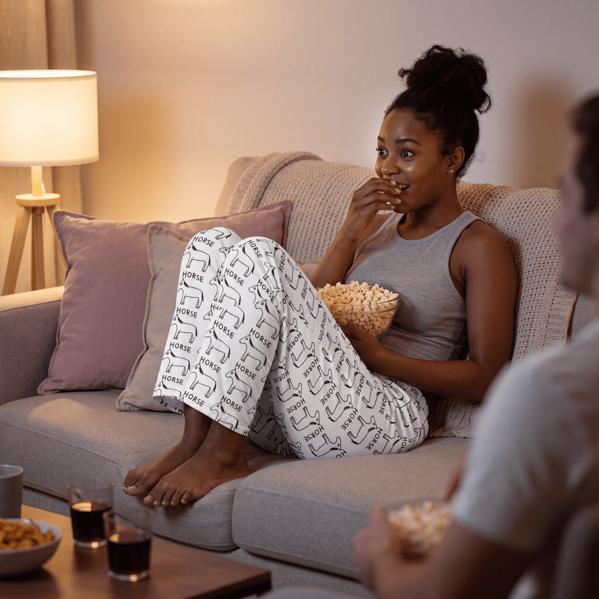 A woman relaxes on a light gray sofa in a living room, wearing white pajama pants with a black geometric pattern and a gray tank top. She holds a bowl of popcorn, with a lamp, pillows, and drinks visible in the cozy scene.