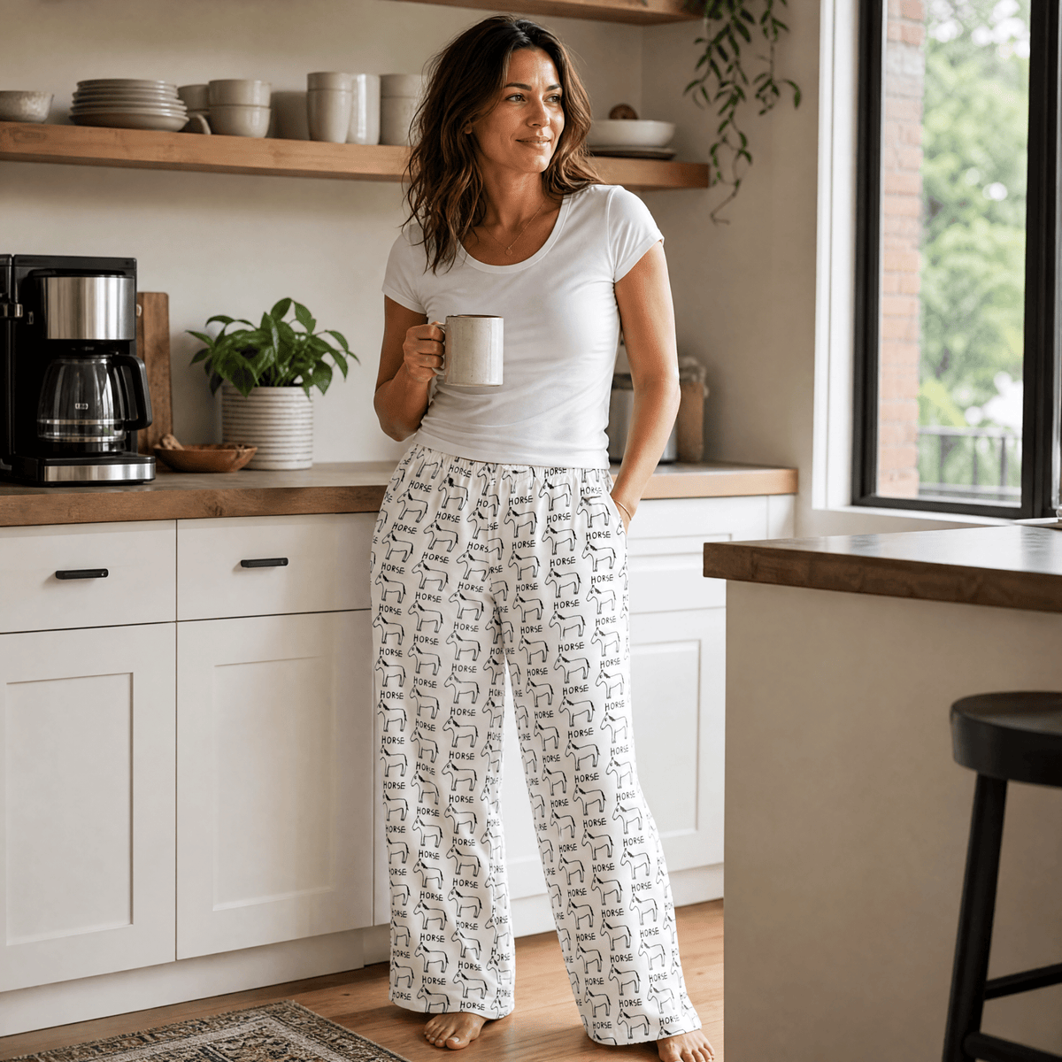 A woman stands in a bright kitchen wearing white pajama pants with a black repeating pattern and a white t-shirt, holding a mug. The background features white cabinets, wooden countertops, and a window.