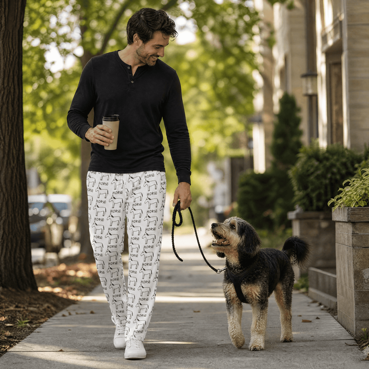 A man walking a small black and tan dog on a sidewalk, wearing a black long-sleeve henley shirt and white pants with a black dog pattern. He is holding a coffee cup and walking in an outdoor residential setting.