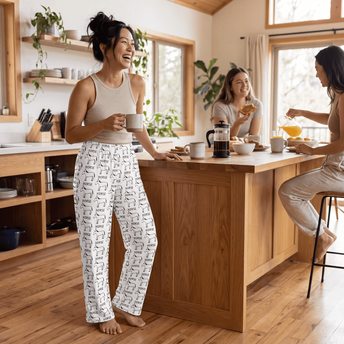 Three women laughing and sharing a breakfast scene in a bright, modern kitchen. The woman in the foreground is wearing beige tank top and white pants with a black geometric pattern, while the others wear neutral-toned casual clothes.