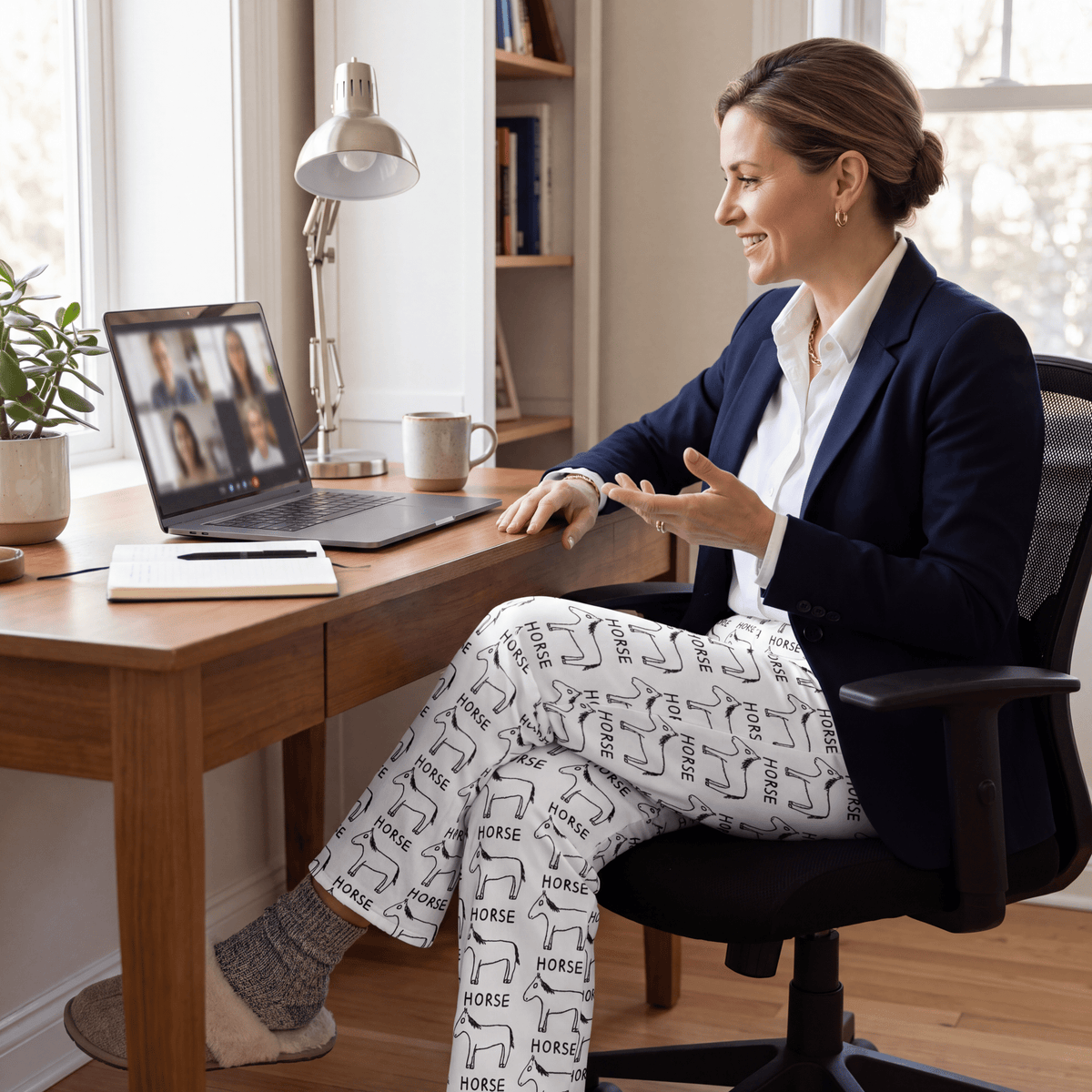 A woman sits in a black office chair at a wooden desk, smiling while talking on a laptop during a video call. She is wearing a navy blazer, white collared shirt, and white pants with a black horse pattern.