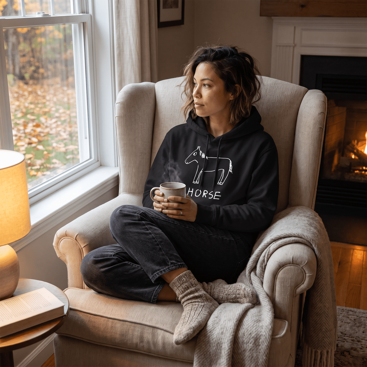A woman sits cross-legged in a beige armchair by a window, holding a mug. She is wearing a black hoodie with a white horse graphic and the word 'HORSE' printed on the front, dark jeans, and knit socks.
