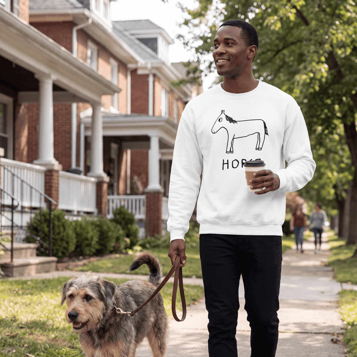 A man walking a dog on a sidewalk in front of brick houses, wearing a white crewneck sweatshirt with a black donkey graphic and the word 'HORSE' printed on the front, holding a coffee cup.