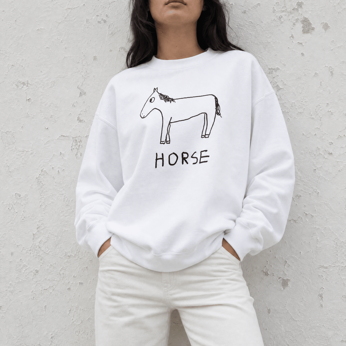 A woman wearing a white crewneck sweatshirt with a black line art drawing of a horse and the word 'HORSE' printed below it, paired with white pants, standing against a textured light gray wall.