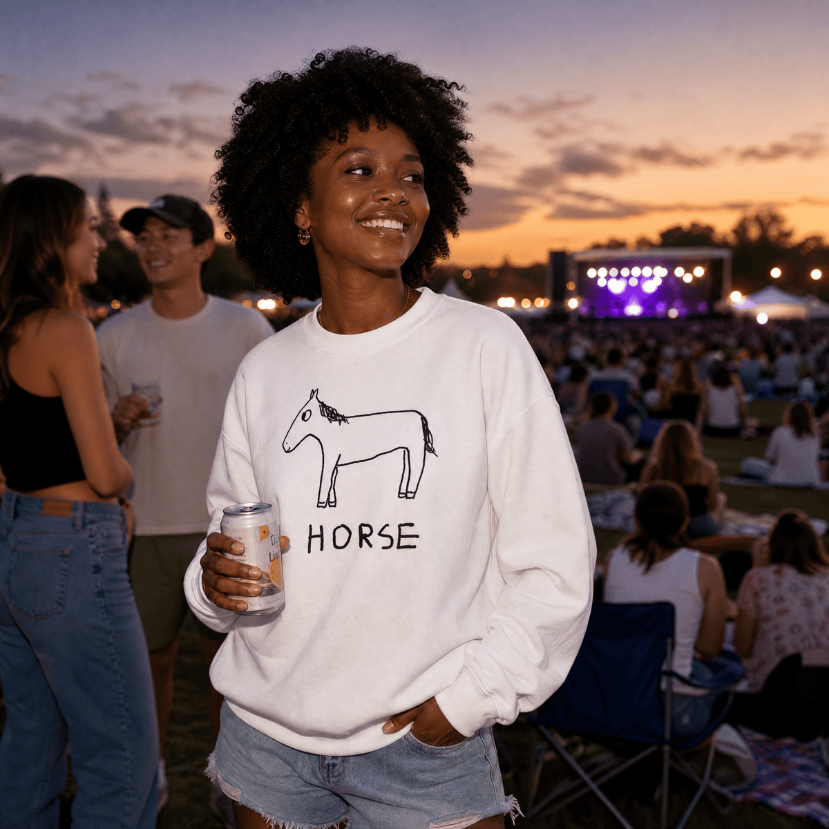 A woman at an outdoor evening concert wears a white crewneck sweatshirt with a black line drawing of a horse and the word 'HORSE' printed on the front. She is holding a beverage can and smiling, with other people and a stage visible in the background.