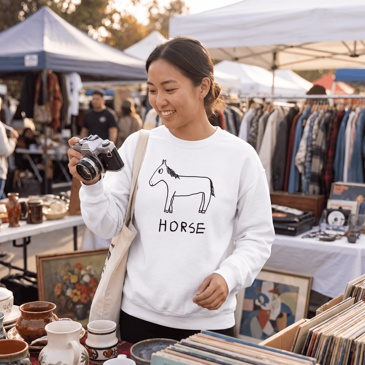 A woman at an outdoor vintage flea market wearing a white crewneck sweatshirt with a black line art horse graphic and the word 'HORSE' printed below it. She is holding a vintage camera and wearing a canvas shoulder bag.
